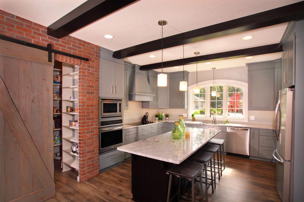 Kitchen with gray painted cabinets, dark stained island and beams and a sliding barn door leading into a pantry